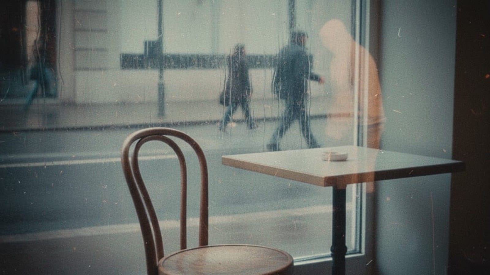 Empty café chair by rain-streaked window with ghostly silhouettes of people walking past outside, shot on vintage film - capturing the space between wanting connection and finding it