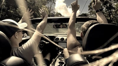 Passengers in a convertible Mustang with hands raised, shot from the back seat, trees overhead.